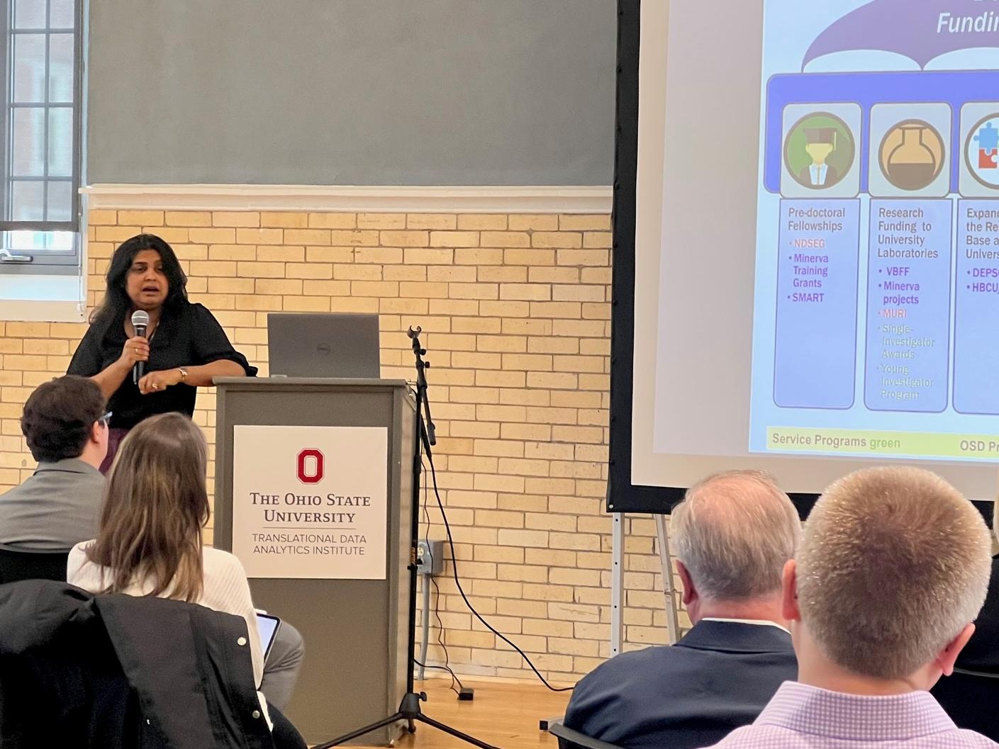 Bindu Nair standing at a podium and holding a microphone speaking to a group of people.