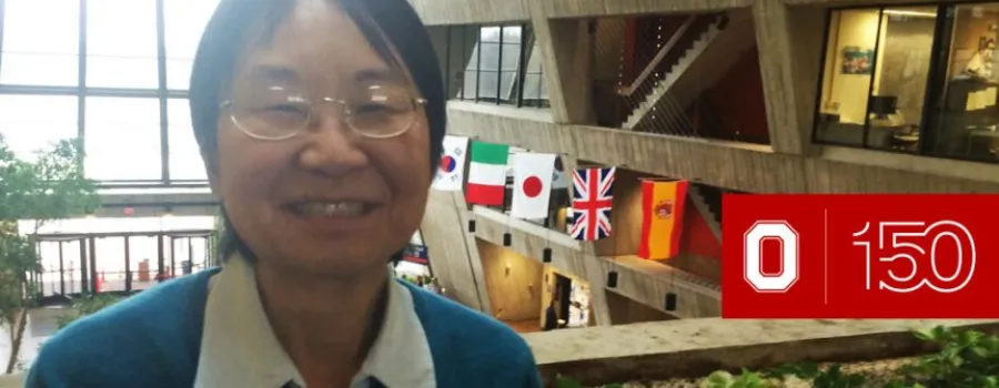 Close up of Junko Shigemitsu wearing glasses sitting on the balcony of an open concept building, International flags to his left, and a windowed wall behind him