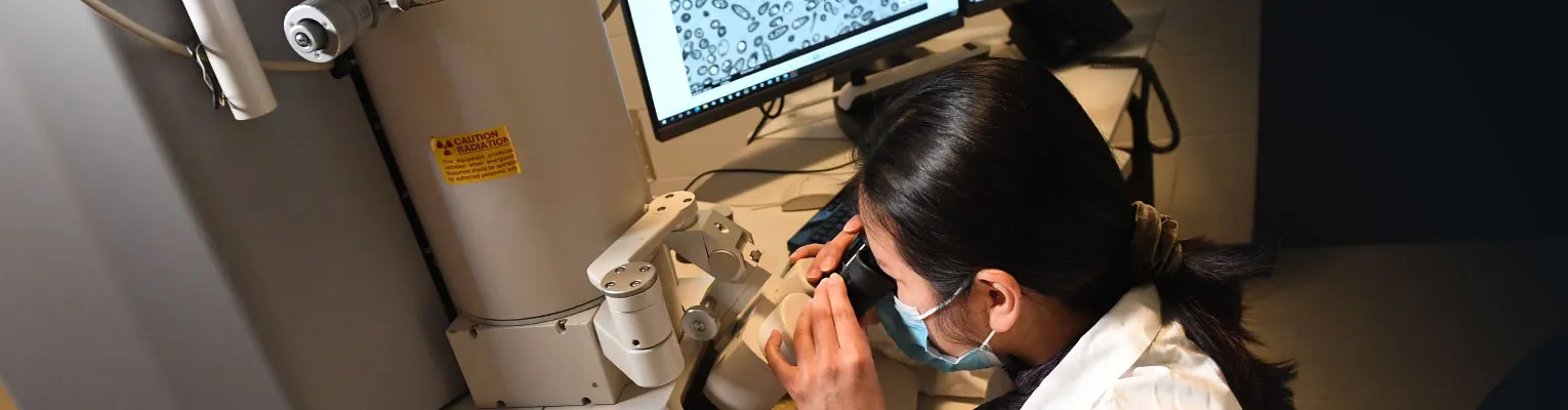 research in lab coat looking into microscope with a monitor display of cells behind the person