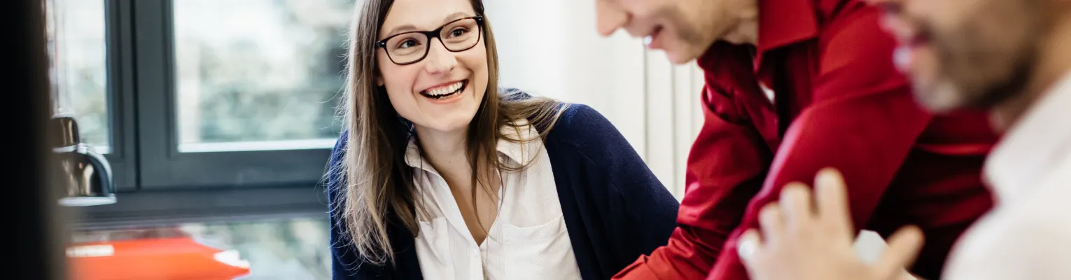 A woman wearing glasses is sitting at a desk in an office smiling at a colleague explaining something to her. The room is bright with big windows in the back and another colleague in the front.