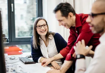 A woman wearing glasses is sitting at a desk in an office smiling at a colleague explaining something to her. The room is bright with big windows in the back and another colleague in the front.