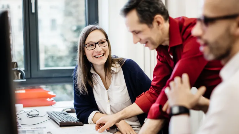 A woman wearing glasses is sitting at a desk in an office smiling at a colleague explaining something to her. The room is bright with big windows in the back and another colleague in the front.