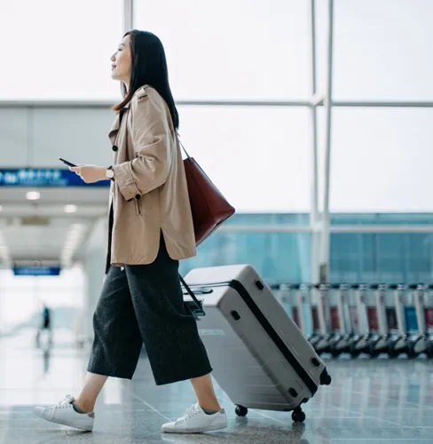 Young Asian woman carrying suitcase and holding smartphone on hand, walking in airport terminal